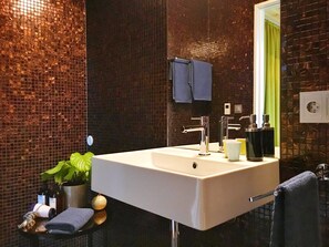 Un baño moderno con un lavamanos grande, dos grifos y un espejo. Hay artículos de tocador en la encimera, un perchero y una planta en maceta.