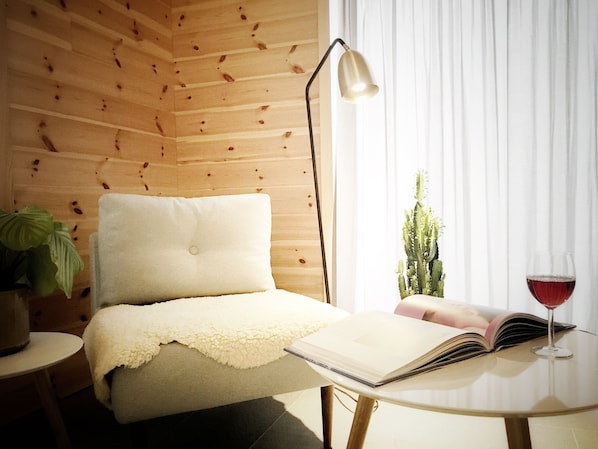 Una habitación acogedora con una pared de madera, un sillón cómodo con cojín blanco, una lámpara de pie, una planta de cactus, una copa de vino tinto y un libro abierto sobre una mesita.