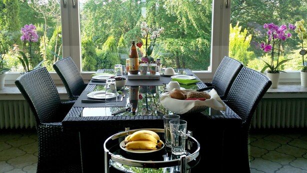 Mesa lista con platos, cubiertos, una botella y un cuenco de fruta, rodeada de sillas, con vistas a la exuberante vegetación y flores a través de grandes ventanales.