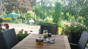 Un área de estar al aire libre con una mesa de madera y sillas de mimbre, rodeada de abundante follaje y flores.