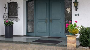 Una puerta doble color turquesa con el número '105' encima, una pequeña ventana con reja decorativa y una planta con flores rosas a la derecha.