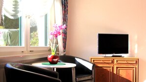 Una sala de estar moderna con un sofá de cuero negro, una mesita redonda con un tazón rojo y flores rosadas, un televisor de pantalla plana sobre un mueble de madera, y un ventanal con cortinas traslúcidas.