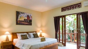A bedroom with a wooden bed, two bedside tables, a nightstand, a painting on the wall, and a view of greenery through large windows. - Alam Pangkung Ubud (Ubud)