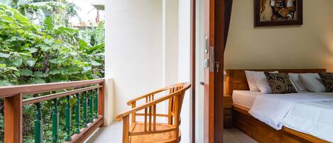 A room with a wooden bed, a wooden chair, and a balcony with a view of greenery.