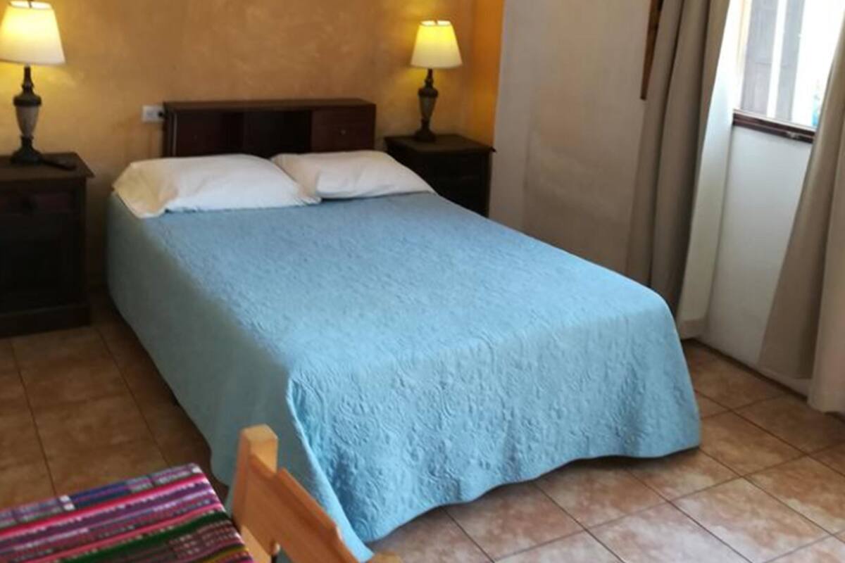 A bedroom with a bed, two bedside lamps, a window with curtains, and a chair with a colorful blanket.