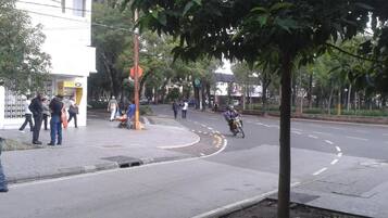 Cena de rua com um edifício à esquerda, uma faixa de pedestres e um motociclista ao longe.
