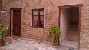 A rustic building with a tiled floor, a wooden door, a window with curtains, and a potted plant.