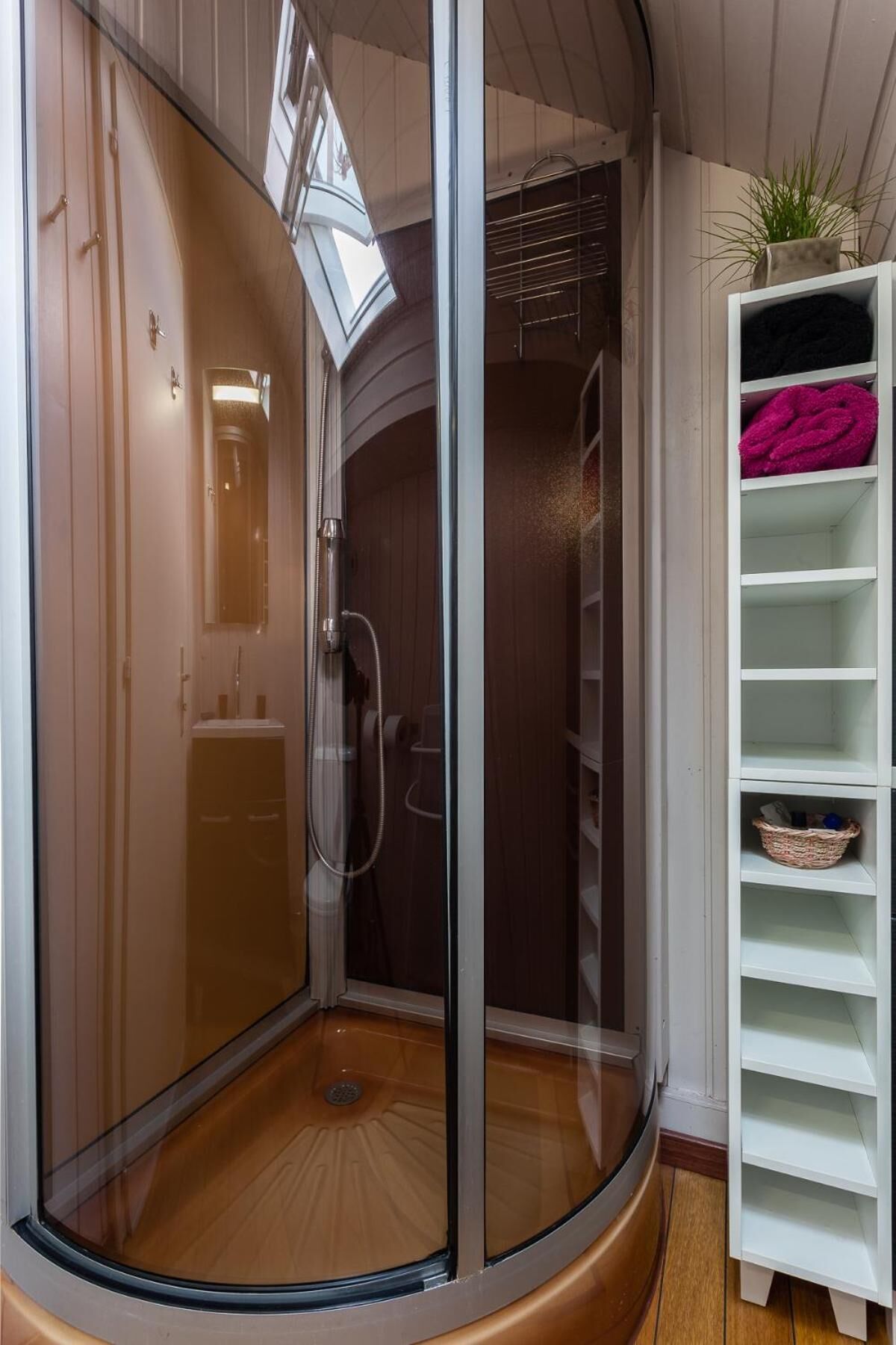 Een douchekamer met glazen wanden, een houten vloer, een wit open kastje met een mand en een potplant.
