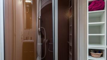 Een douchekamer met glazen wanden, een houten vloer, een wit open kastje met een mand en een potplant.