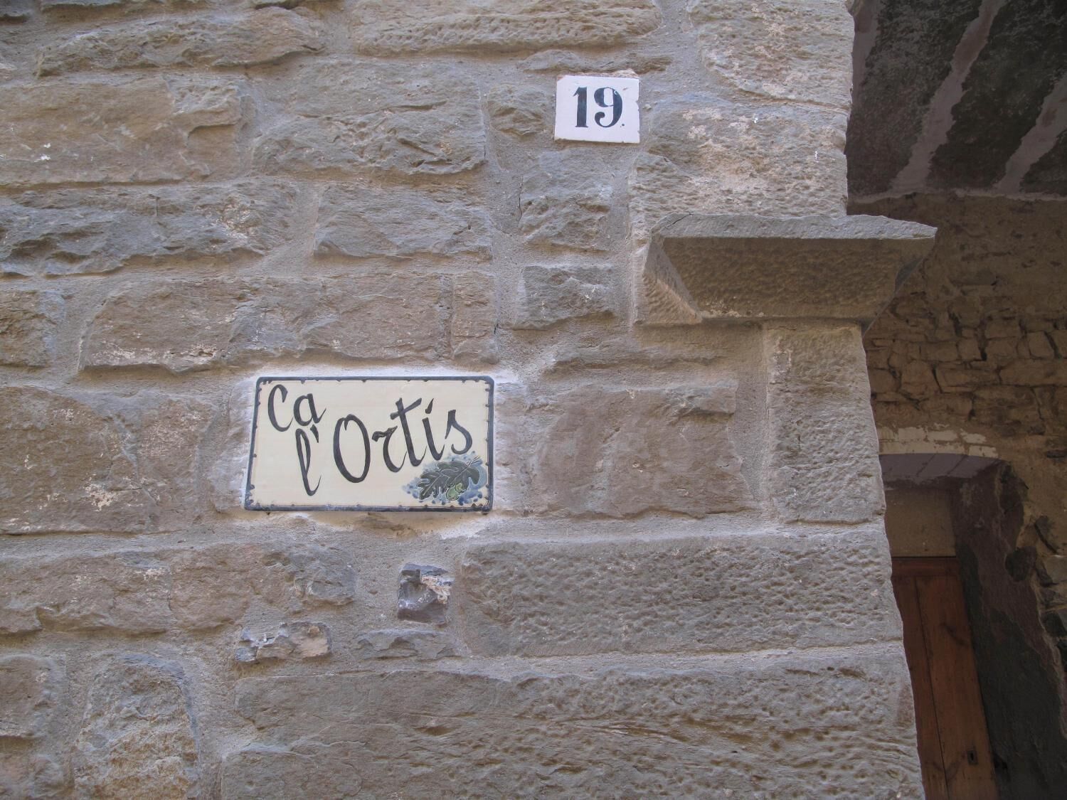 A stone wall with a house number sign '19' and a door plaque reading 'Ca l'Ortis'.