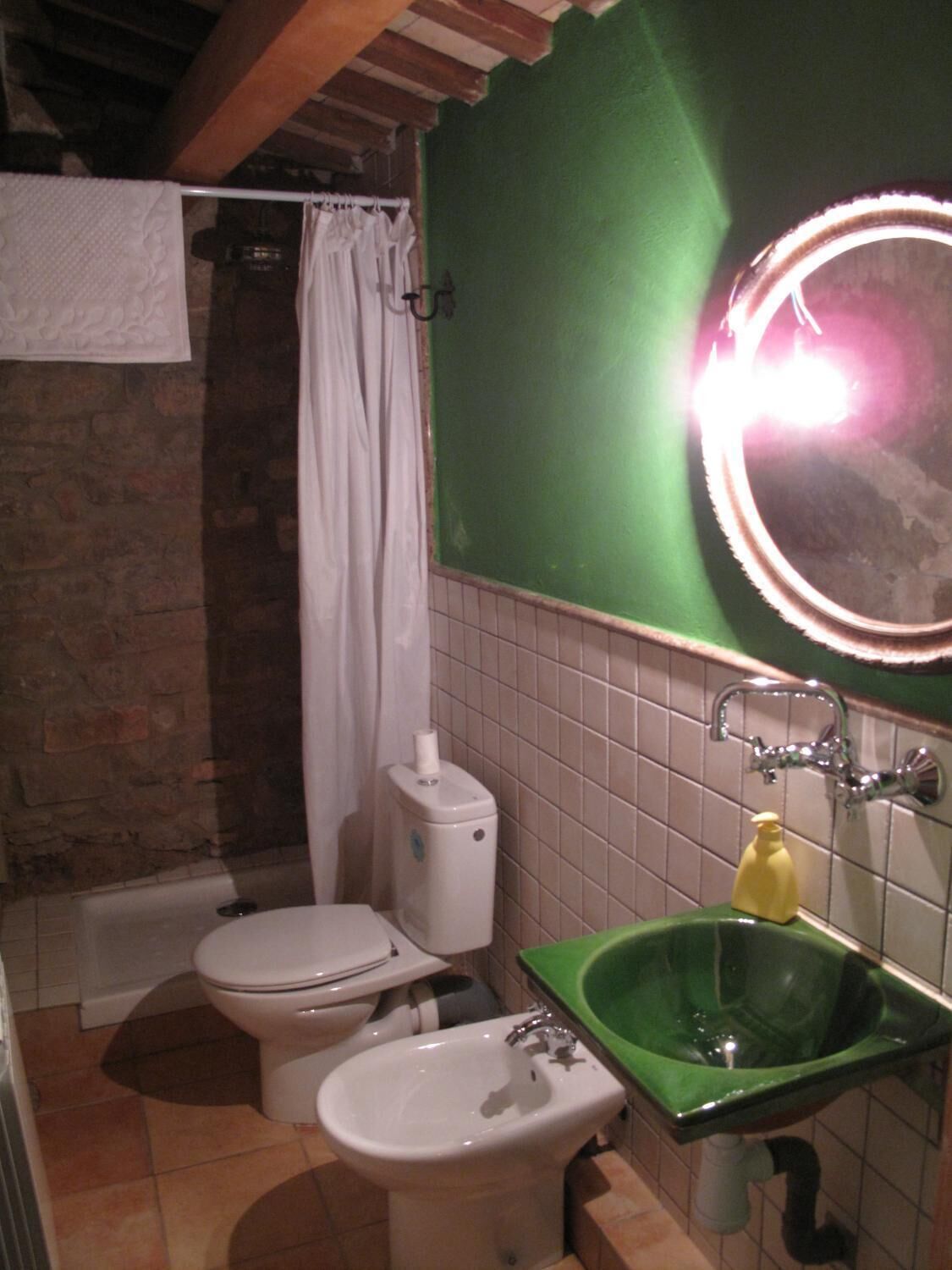 A bathroom with a toilet, bidet, green sink, and a mirror with a light.
