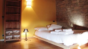 A bedroom with a wooden bed, a nightstand with a lamp, a wooden shelf with baskets, and a stone wall.