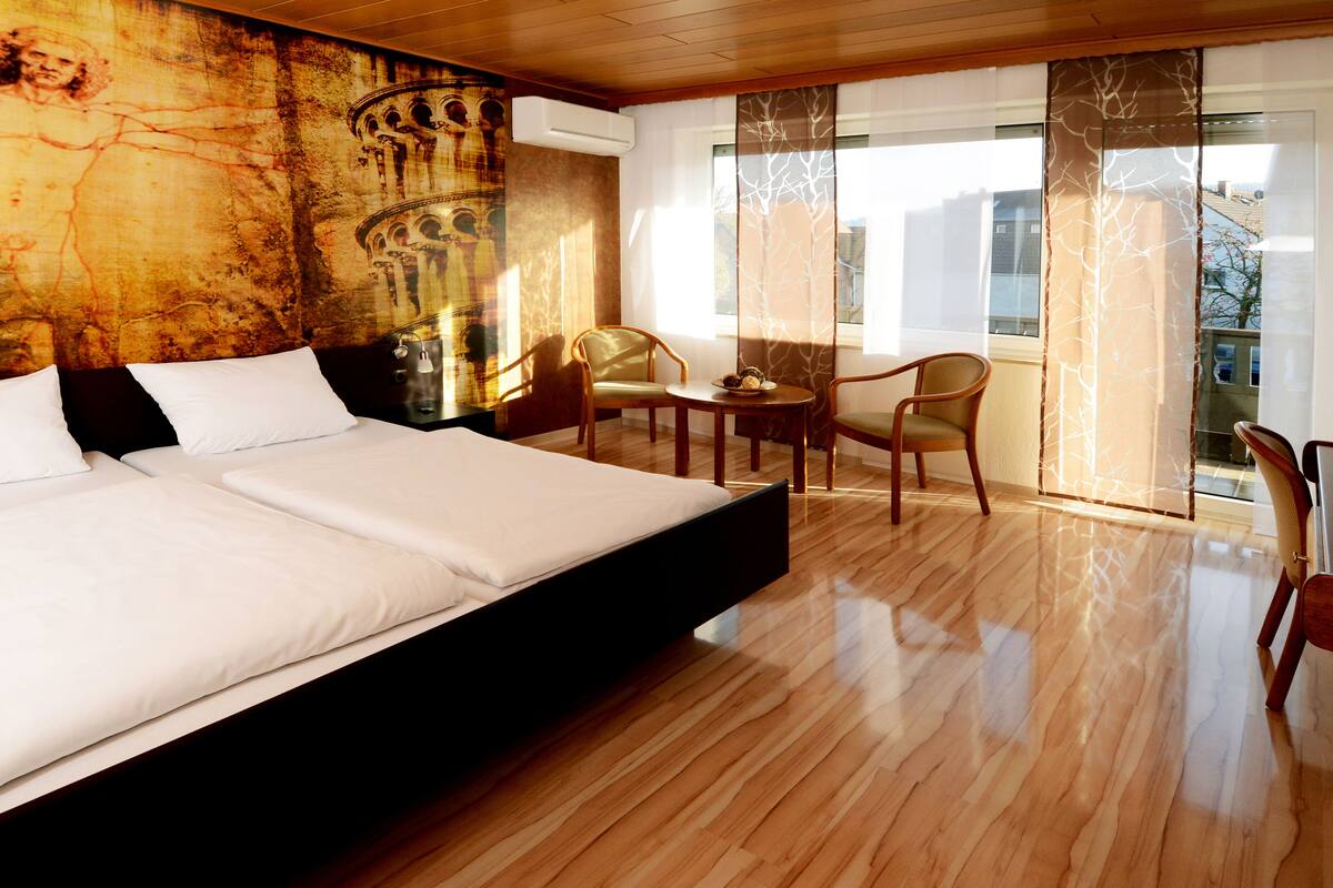A hotel room with a large bed, wooden flooring, a dining area with chairs, and a view through the window.