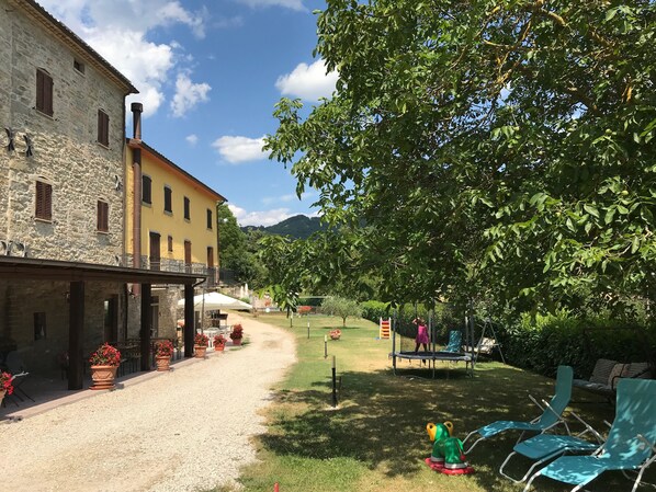 A courtyard with a stone building, a yellow building, and a playground with a trampoline and swings. - Agriturismo Cailuca (Pietralunga)