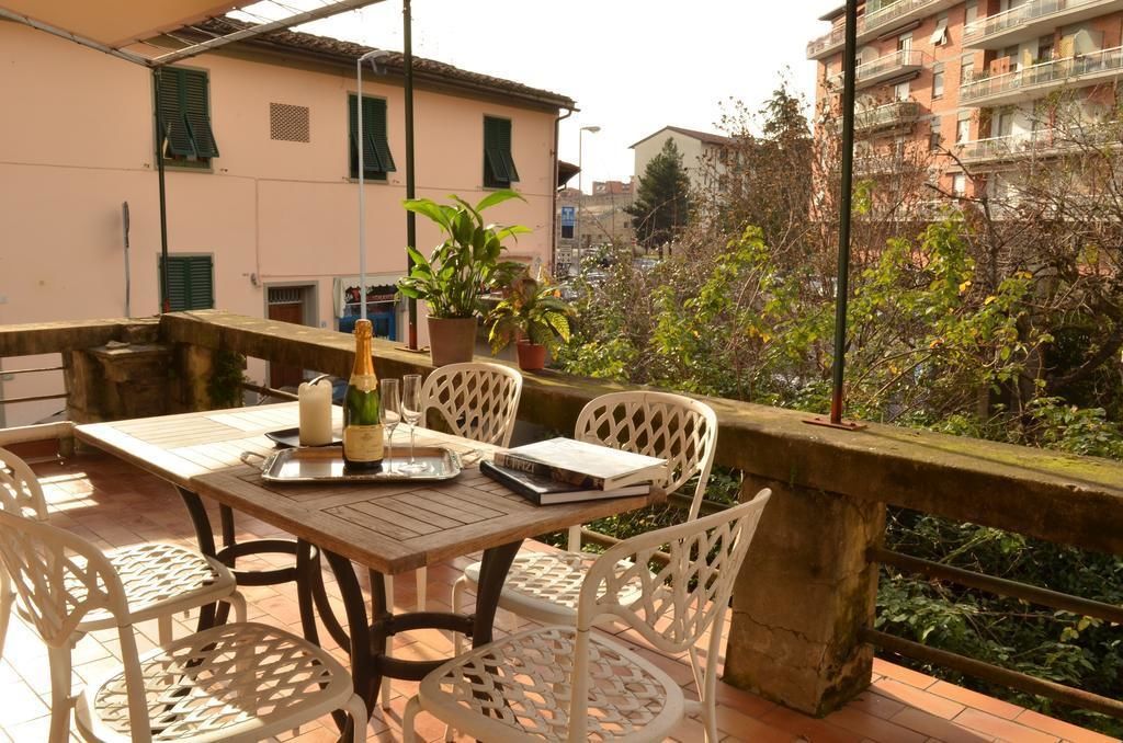 A terrace with a table set for two, surrounded by white chairs, with a view of buildings and greenery in the background.