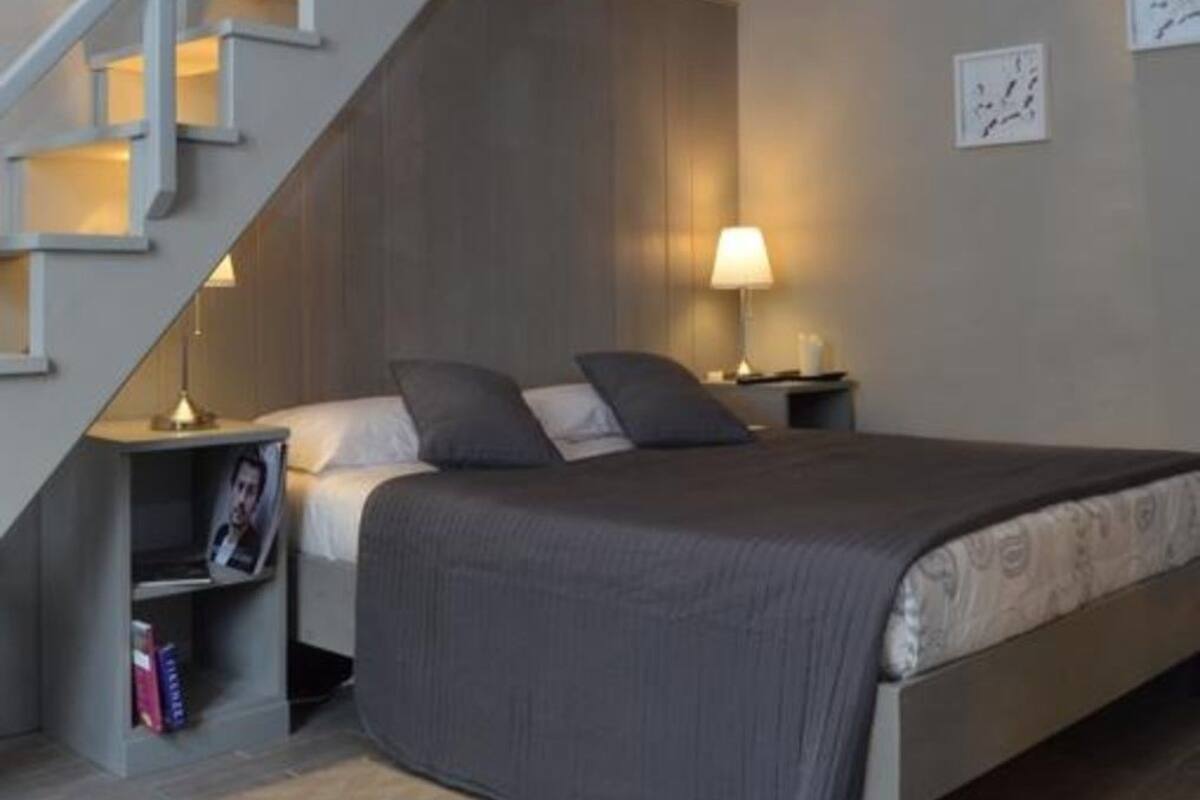 Un dormitorio con cama, mesita de noche, lámpara, un estante pequeño con libros y una escalera que lleva a un nivel superior.