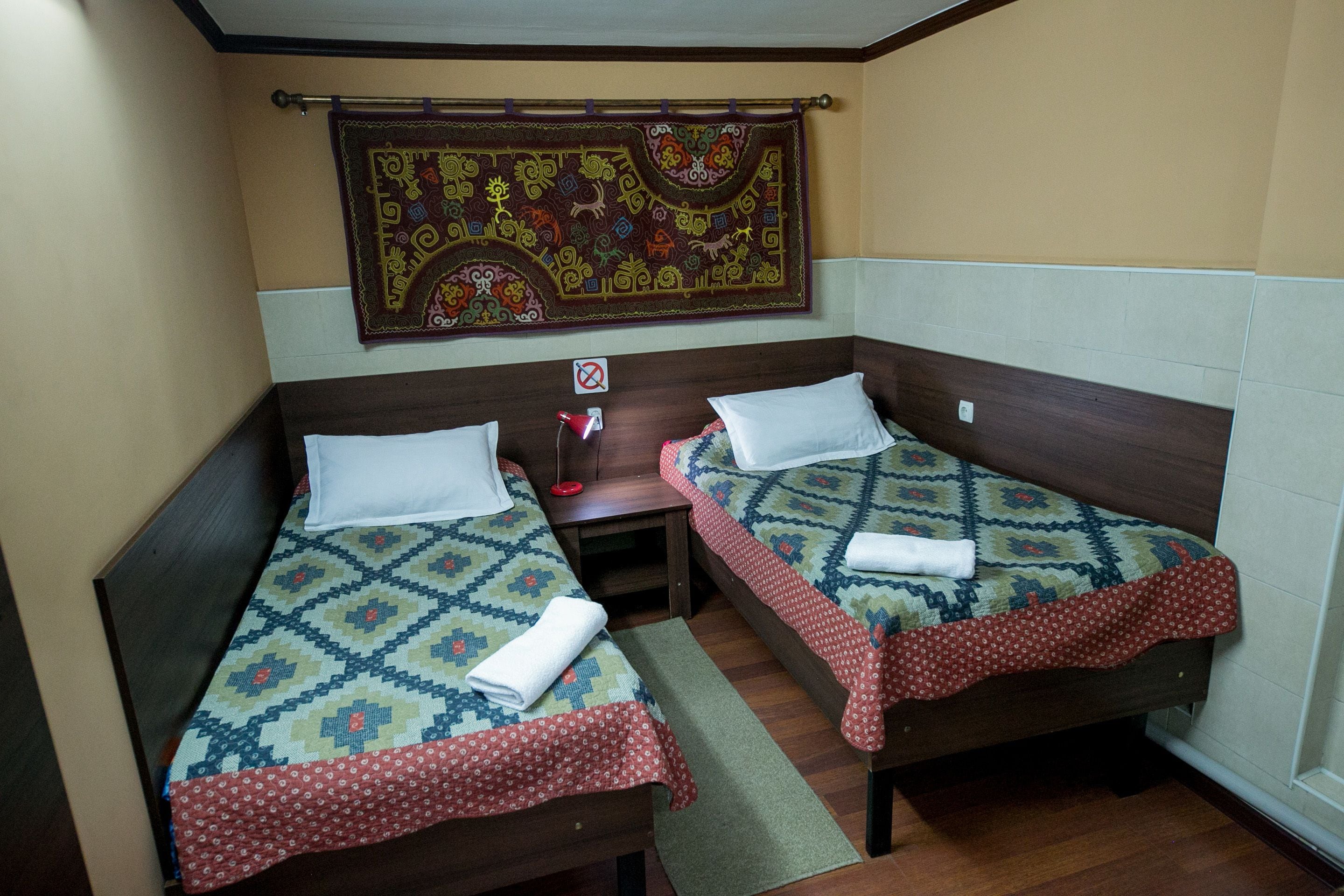 Une petite chambre avec deux lits simples, une table de chevet, une tapisserie murale et une petite table sur laquelle se trouve une lampe rouge.