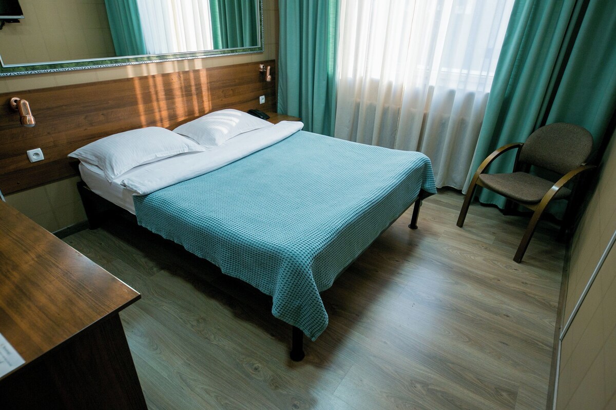 A hotel room with a wooden headboard, a bed with a blue bedspread, a wooden desk, a chair, and a window with curtains.