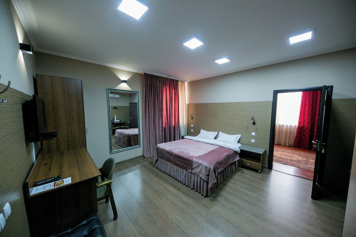 Quarto de hotel com uma secretária de madeira, uma cama com cabeceira, um espelho e uma porta que dá para outro quarto.