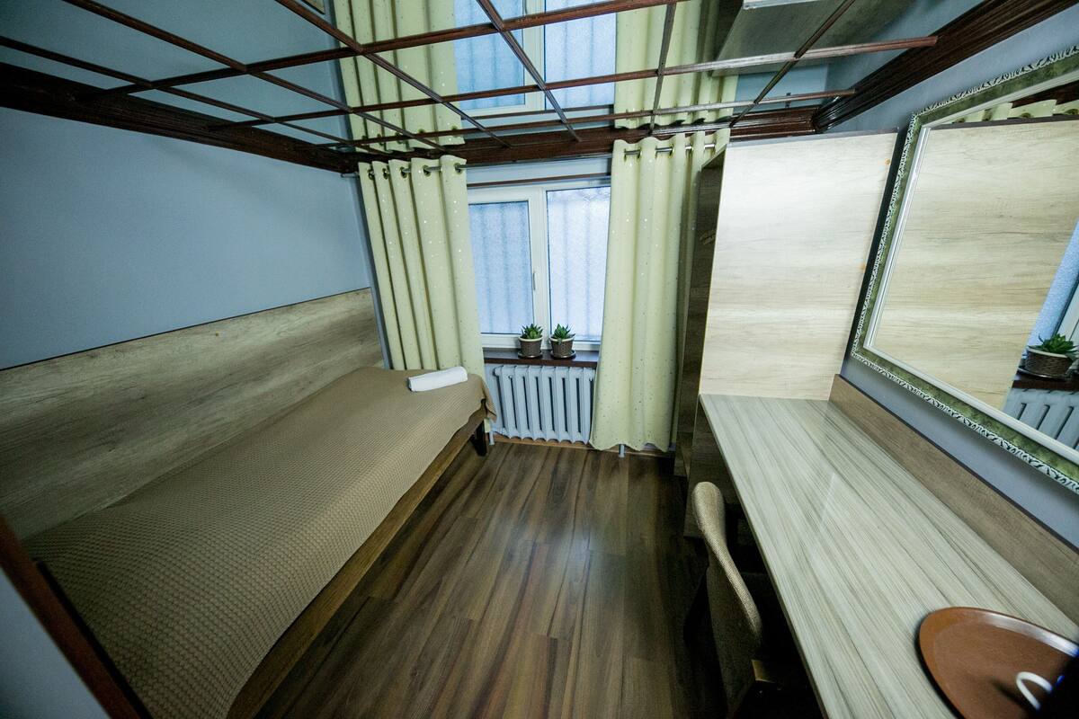 Una habitación pequeña con suelo de madera, una cama, una silla, un ventanal con cortinas y un radiador.
