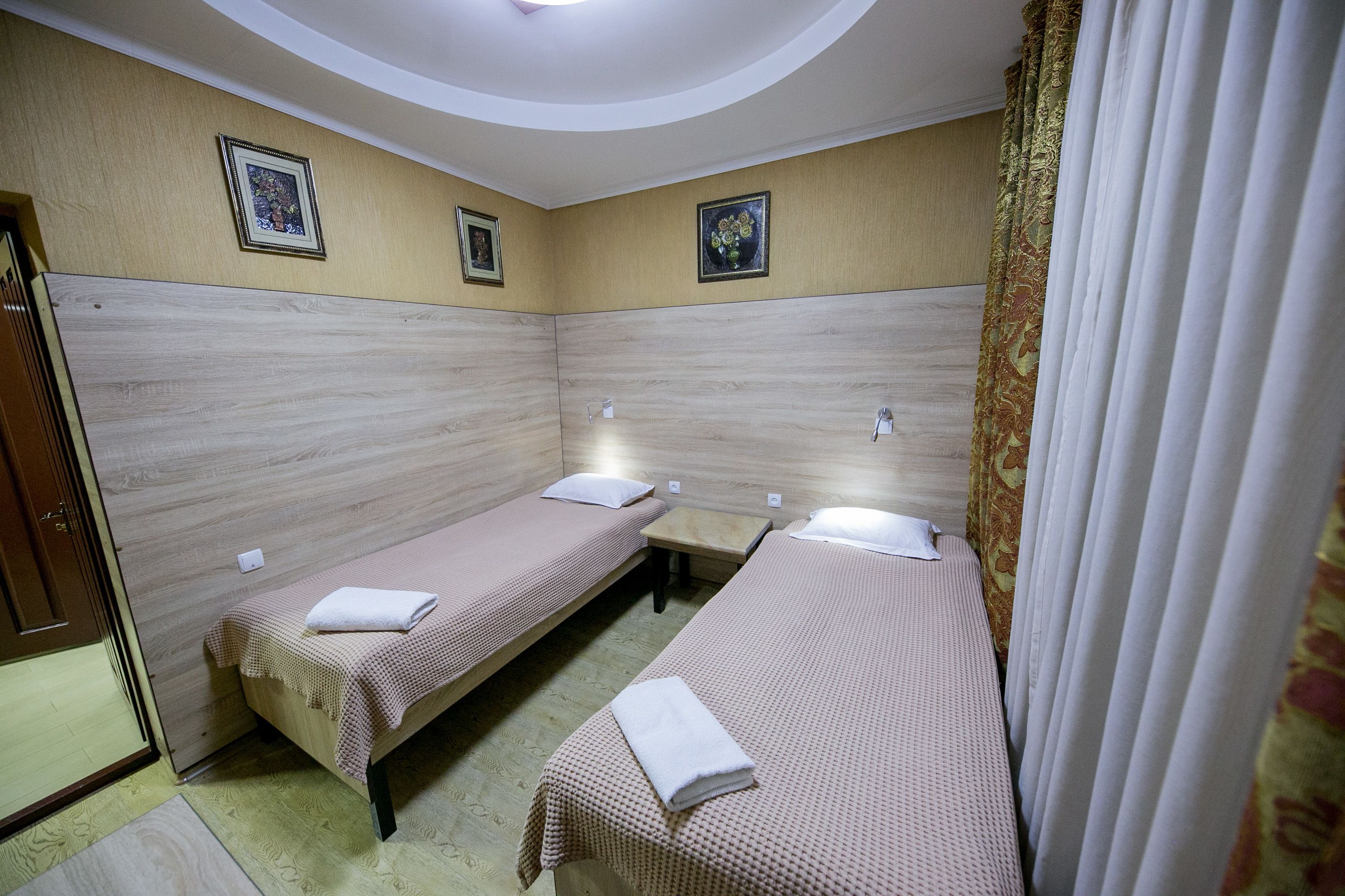A room with two single beds, each with a pillow and a folded towel. The walls are beige with a wood-like texture, and there are two framed pictures on the wall.