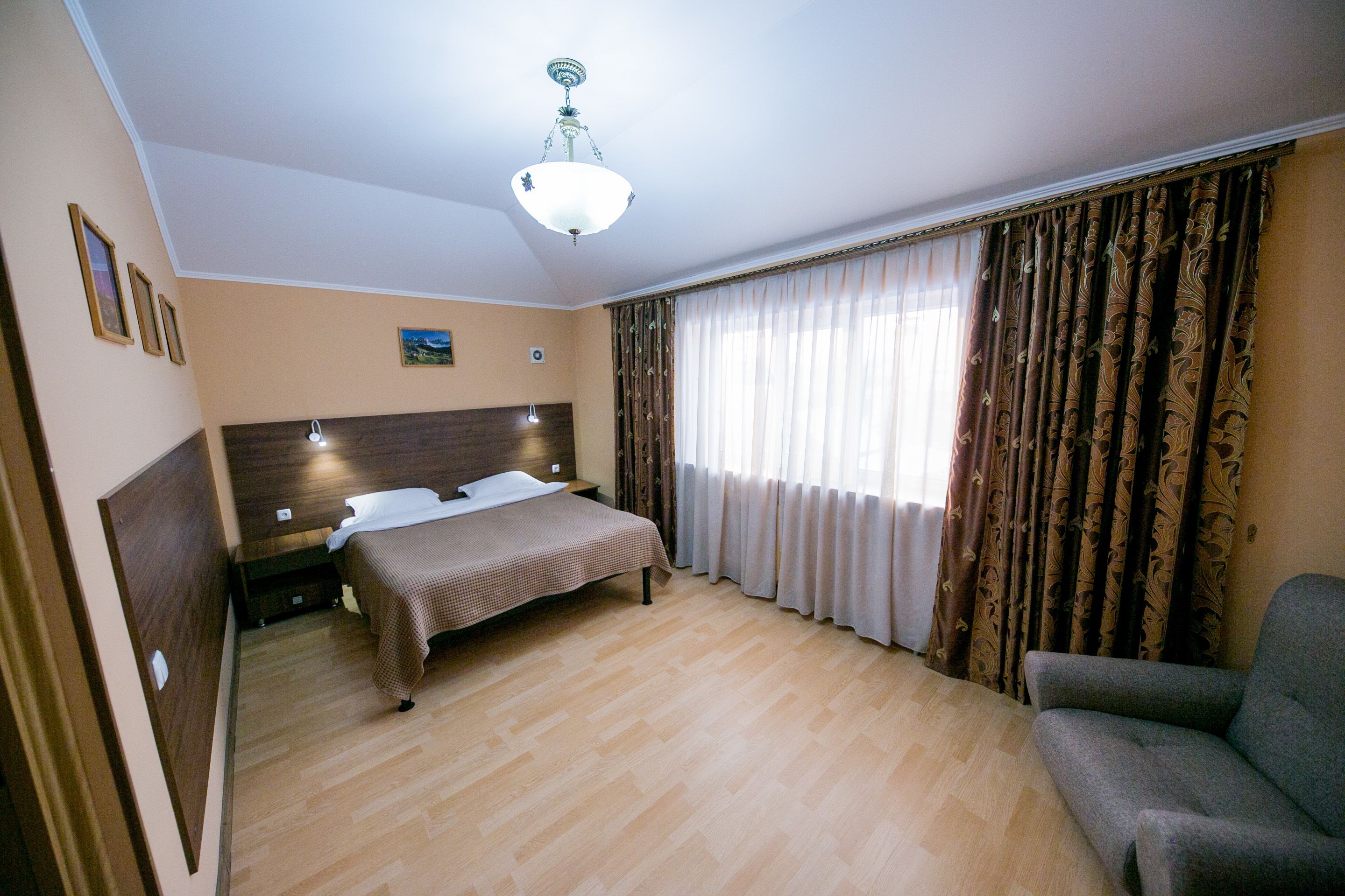 Ein Hotelzimmer mit einem Bett, einem Sessel, einem Nachttisch, einem Holz-Kopfstück, einer Deckenleuchte und Vorhängen.