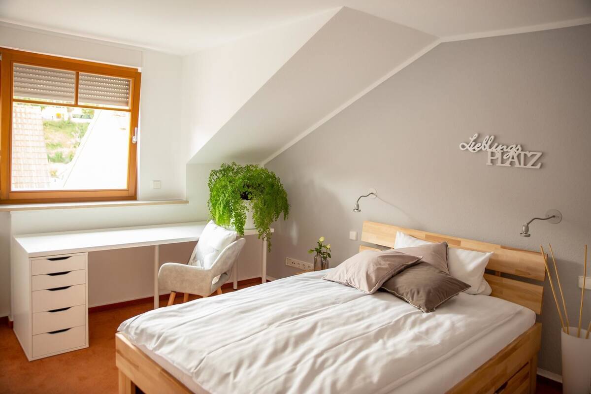 A bedroom with a sloped ceiling, a desk with drawers, a chair, a bed with pillows, a hanging plant, and a wall-mounted light fixture.