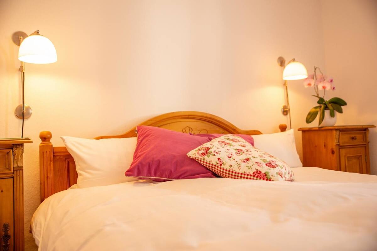 A neatly made bed with white and pink bedding, two wall-mounted lamps, and a wooden nightstand with a plant.