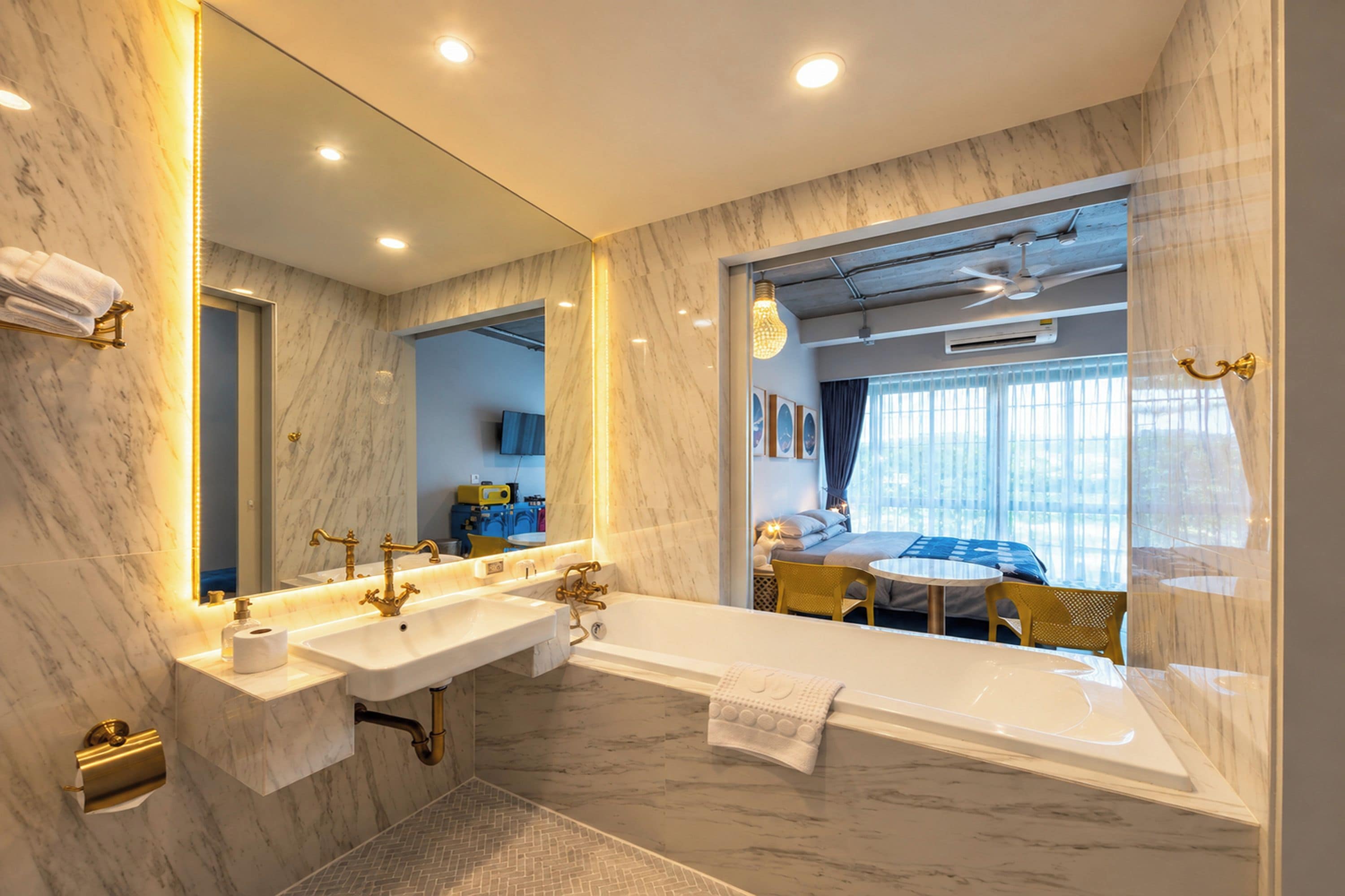Une salle de bain moderne dotée d’un grand miroir, de deux vasques, d’une baignoire et d’une chambre à coucher visible, avec un lit et une fenêtre.
