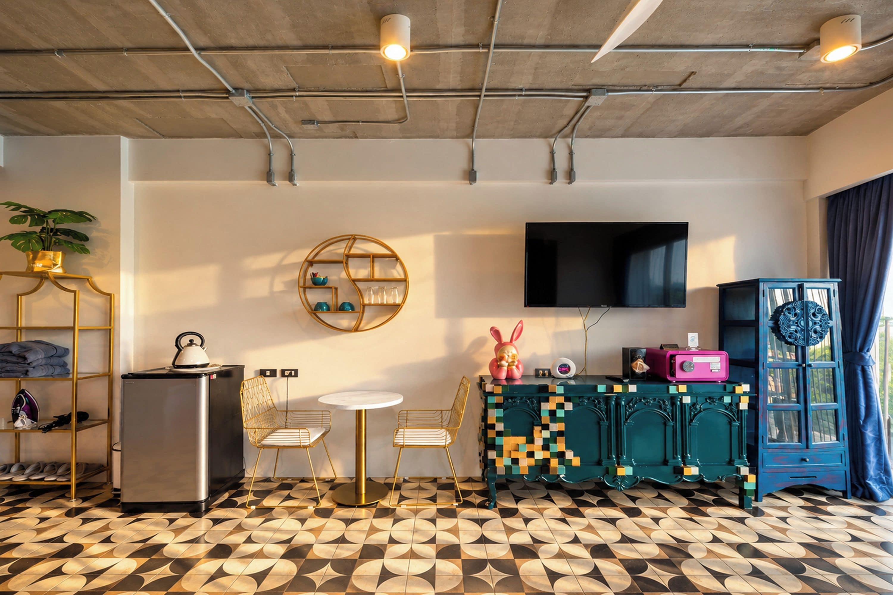 A modern living room with a flat-screen TV, a round wooden shelf, a small table with chairs, a refrigerator, and a patterned floor.