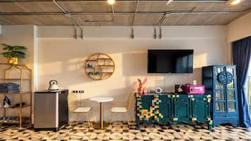 A modern living room with a flat-screen TV, a round wooden shelf, a small table with chairs, a refrigerator, and a patterned floor.