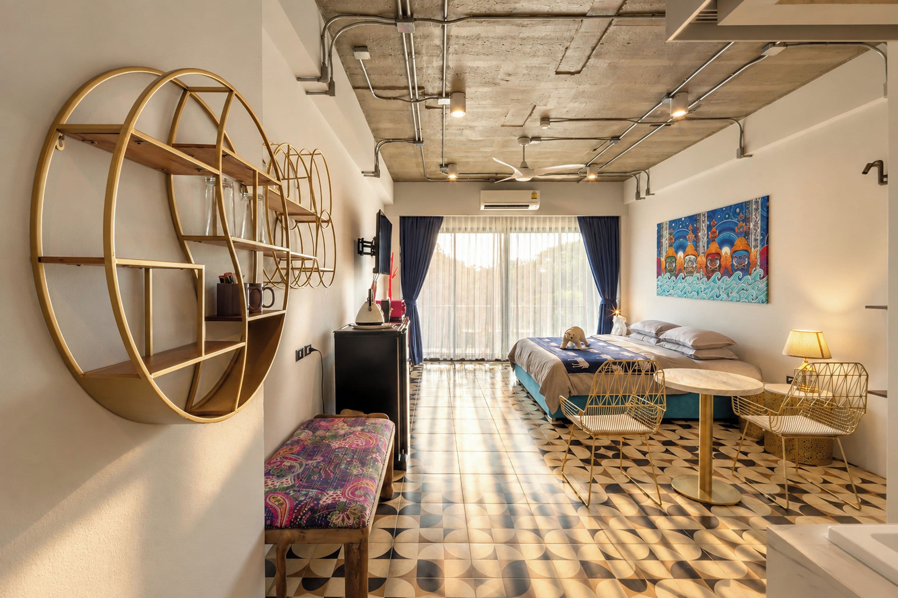 Habitació d'hotel moderna amb un sòl de disseny geomètric, una prestatgeria circular gran, un llit amb un capçal colorit, una taula rodona petita amb una làmpada i un quadre penjat a la paret.