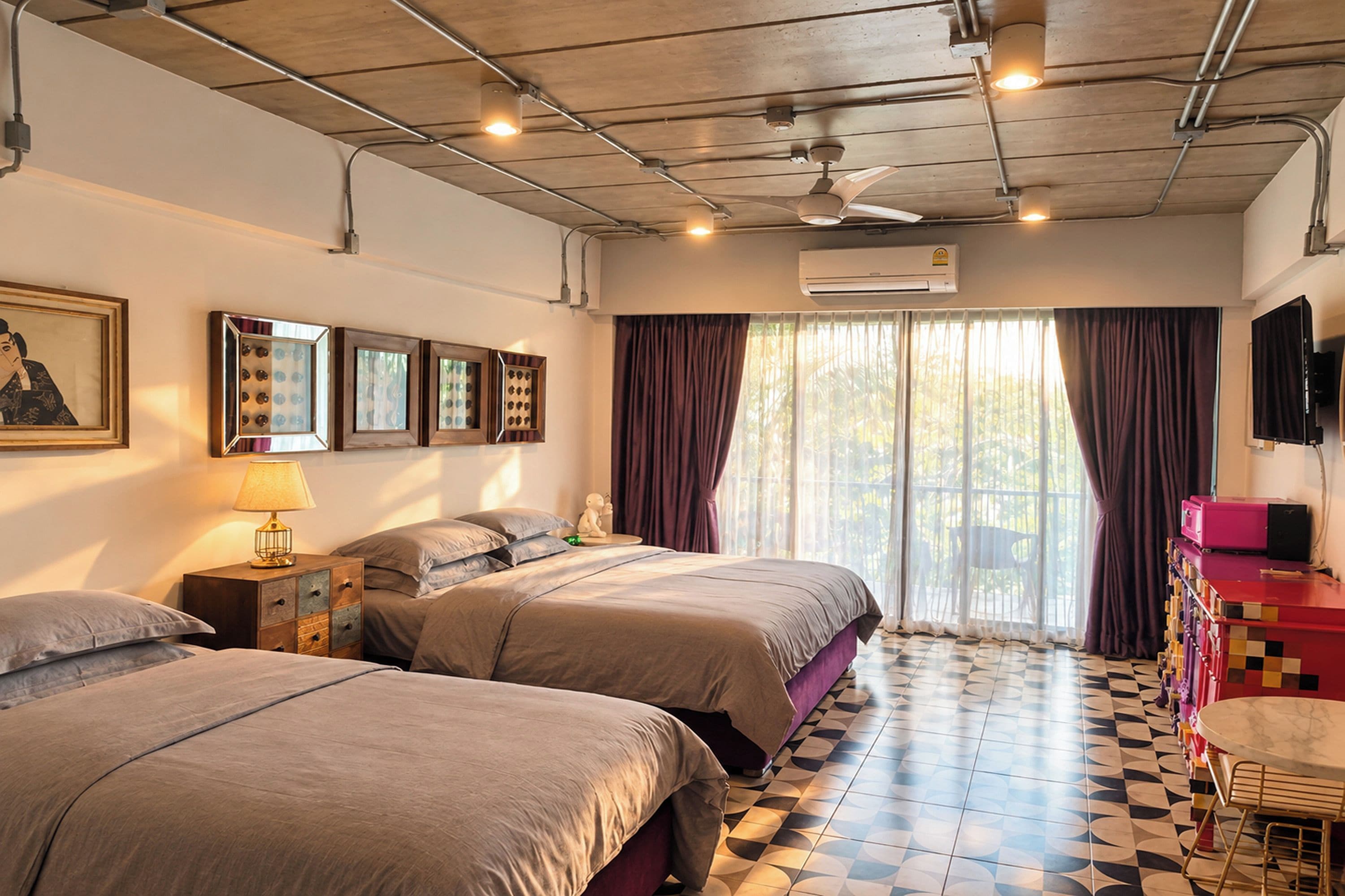 A hotel room with two beds, a ceiling fan, an air conditioning unit, a television, and a window with curtains.