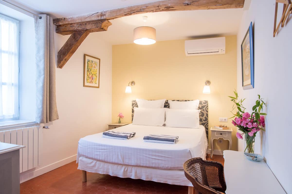 Une chambre à coucher comprenant un lit, des tables de chevet, une fenêtre avec des rideaux, une lampe suspendue, un appareil de climatisation et un tableau encadré au mur.