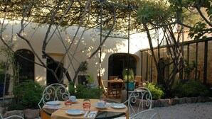 Un espace de restauration en plein air avec une table ronde et des chaises en métal blanc, entouré de verdure et bordé par un bâtiment aux fenêtres en arc de cercle.