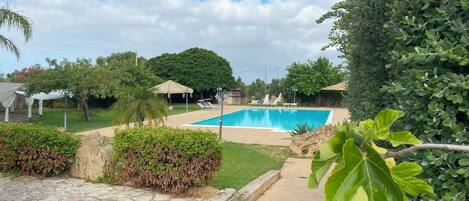 Piscina com um caminho de pedra que a leva, rodeada por vegetação e uma grande árvore.