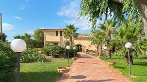 A pathway leading to a building with a tiled entrance, surrounded by potted plants and palm trees. - Agriturismo Le Arcate (Marsala)