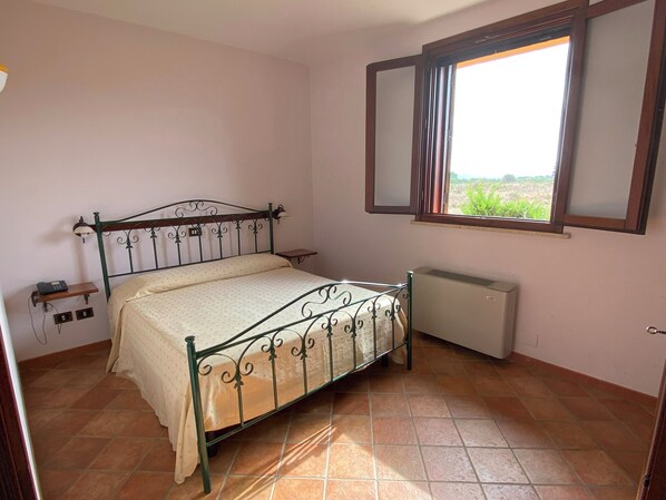Une chambre avec un lit en métal, une table de chevet, un radiateur et une fenêtre donnant sur un paysage verdoyant.