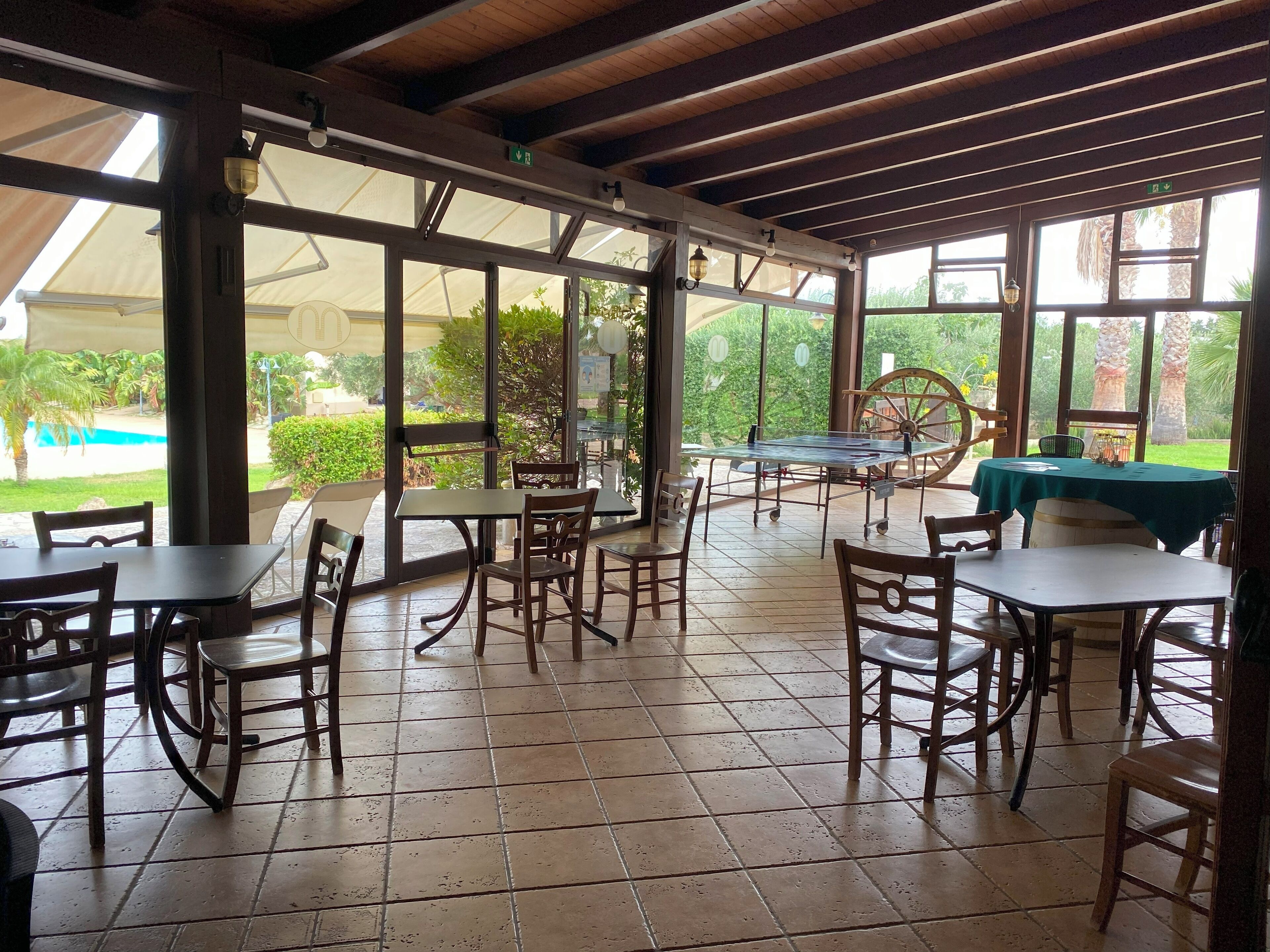 Un'area patio esterna con tavoli e sedie, ampie finestre e vista su una piscina e vegetazione.