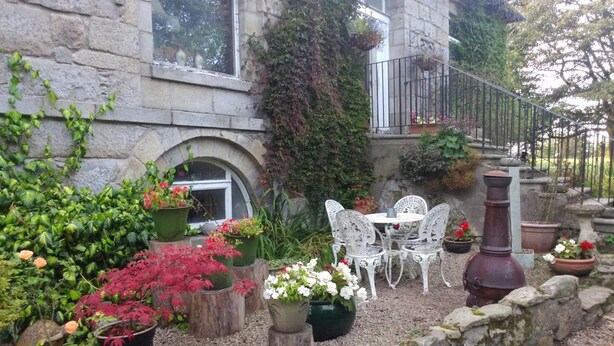 A stone building with a garden patio featuring white metal chairs and tables, surrounded by various potted plants and a stone wall. - Disblair House (Aberdeen)