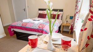 A neatly arranged breakfast table with plates, cups, and cutlery, a vase with flowers, and a bed with a floral bedspread in the background.