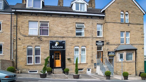 A two-story building with a 'Snooze' sign, indicating a bed and breakfast. There is a 'Budget Rooms' sign on the right. The building has a brick facade with multiple windows and a small porch area with potted plants.