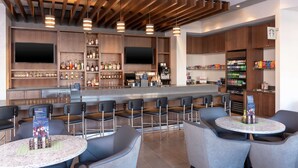 A modern bar area with wooden ceiling beams, multiple seating options including bar stools and round tables, and a variety of beverages and snacks available. - Hyatt Place Detroit Royal Oak (Royal Oak)