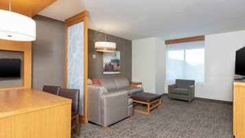A hotel room with a wooden dining table, a gray sofa, a gray armchair, a wooden bench, a TV mounted on the wall, and a window with a view of mountains.