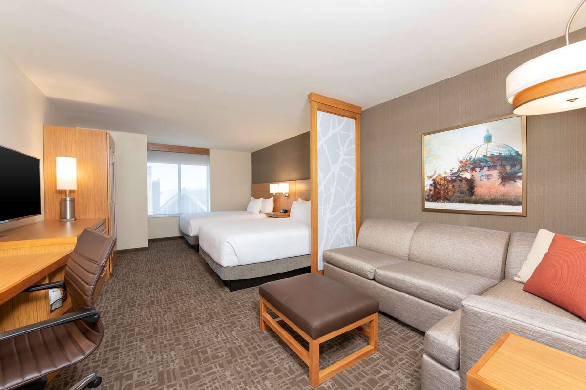 A hotel room with a bed, a sofa, a desk with a chair, a TV, and a painting on the wall.