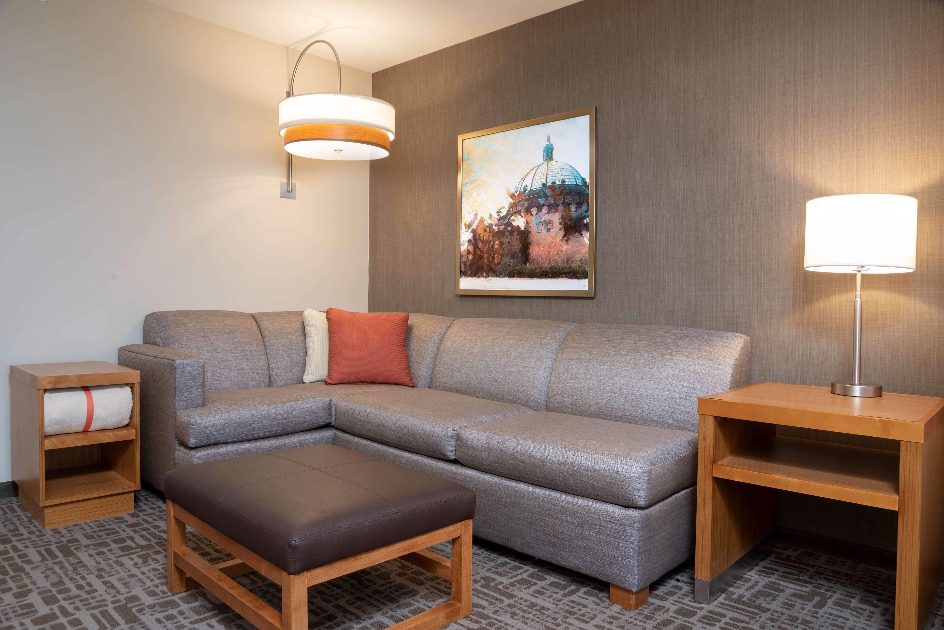 A modern hotel room with a grey sectional sofa, a wooden nightstand, a small wooden table, and a floor lamp.