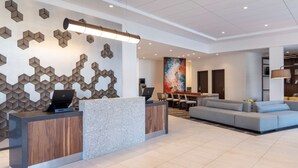A modern reception area with a wooden reception desk, a grey sofa, and a colorful abstract painting on the wall. - Hyatt Place Detroit Royal Oak (Royal Oak)