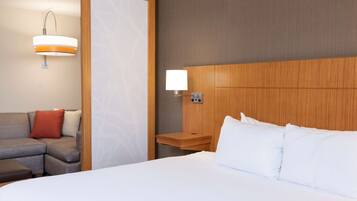 A hotel room with a large bed, a wooden headboard, a nightstand with a lamp, a sofa with cushions, and a wall-mounted light fixture.