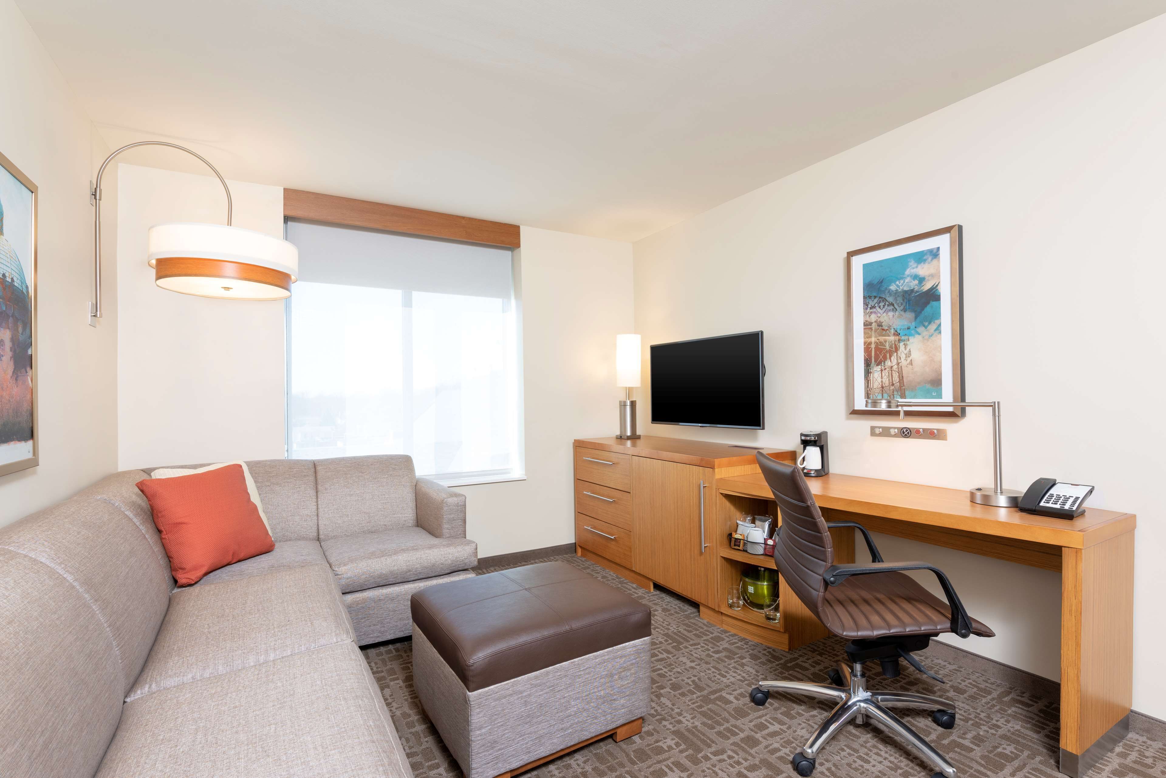 A hotel room with a sofa, ottoman, desk, chair, TV, and artwork on the wall.