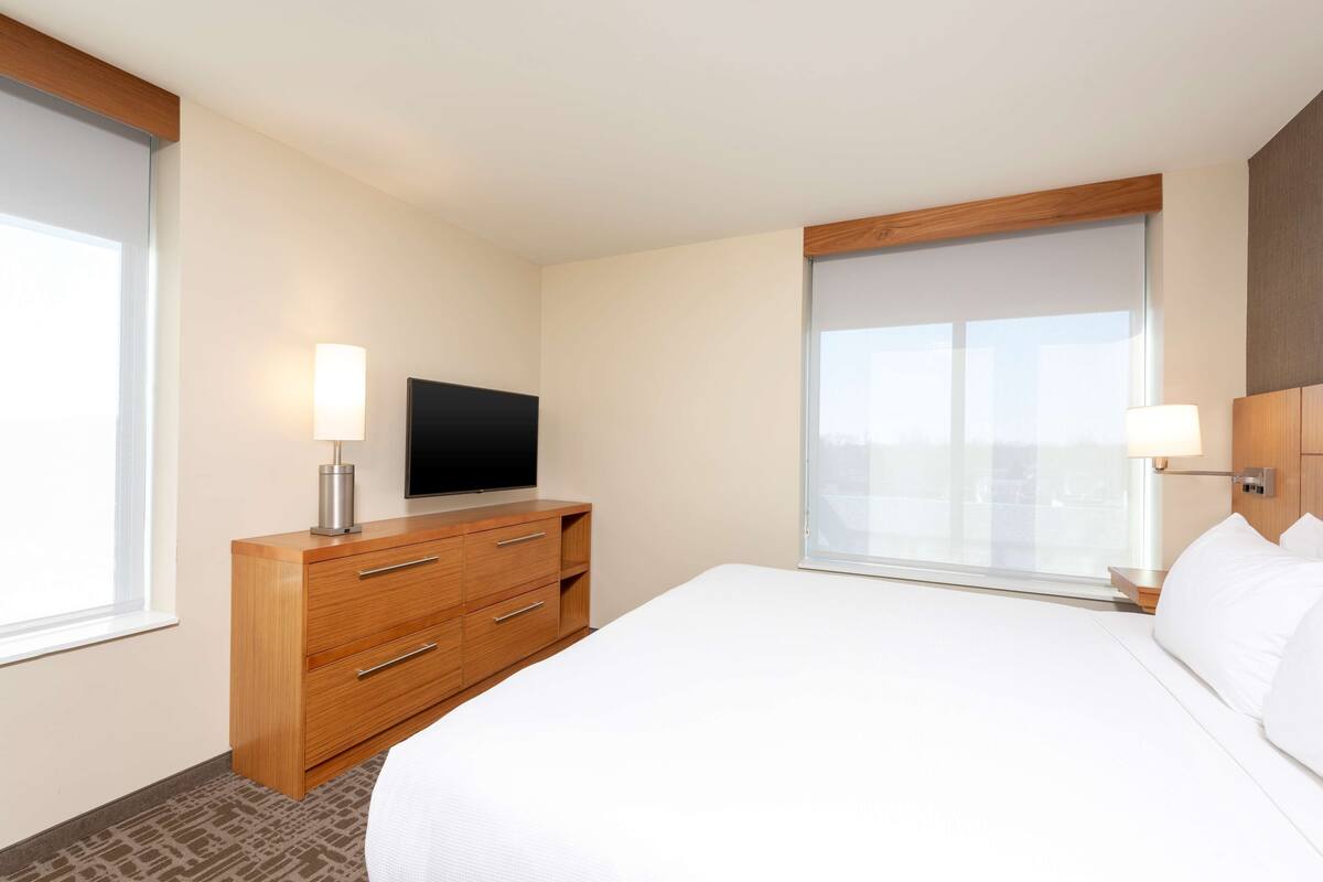 A hotel room with a large bed, wooden dresser, flat-screen TV, and a window with a city view.